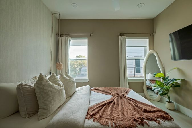 Cozy neutral bedroom — plush white pillows on a large bed with a rust-knit throw, two sunlit windows, oval full-length mirror, potted plant and wall-mounted TV.