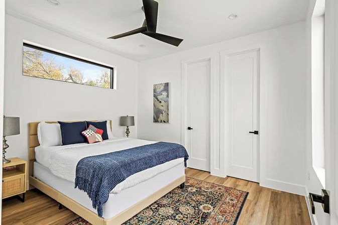 Bright modern bedroom interior with upholstered bed, navy throw and star pillow, wood nightstands with lamps, patterned area rug, long horizontal window and black ceiling fan