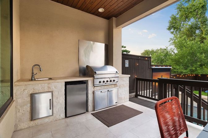 Covered backyard patio outdoor kitchen on a raised deck with stainless-steel grill, mini fridge, sink and stone countertop, red woven chair and string lights overlooking trees and a pool.
