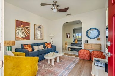 Cheerful modern living room with a blue sofa and patterned pillows, mustard yellow accent chair, round white coffee table on a pink patterned rug, hardwood floors, arched doorway, ceiling fan and red front door visible.