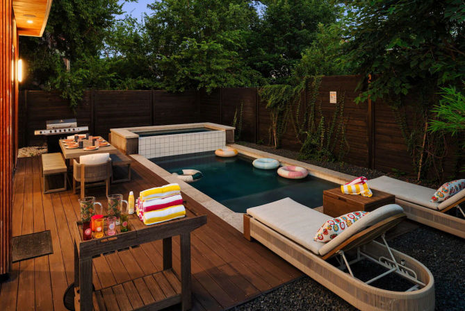 Cozy backyard pool patio at dusk with a small tiled plunge pool and raised hot tub, wooden deck with lounge chairs and colorful float rings, dining table set by a barbecue grill, stacked striped towels and leafy privacy fence.