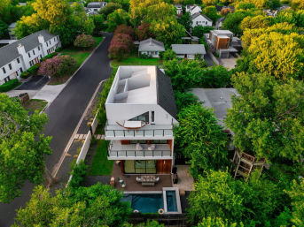 Drone aerial view of a modern three-story suburban home with rooftop terrace, glass balconies and a backyard pool surrounded by lush trees.