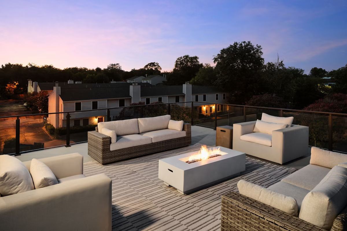 Cozy rooftop patio at twilight with modern wicker sofas and cushioned armchairs around a lit rectangular fire pit, glass railing overlooking suburban townhomes.