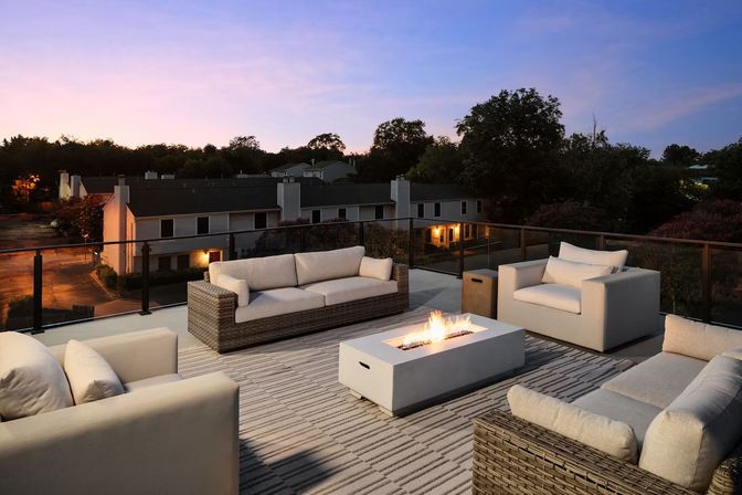 Cozy rooftop patio at twilight with modern wicker sofas and cushioned armchairs around a lit rectangular fire pit, glass railing overlooking suburban townhomes.