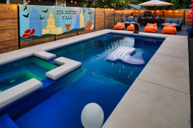 Cozy Austin backyard pool at twilight with connected spa, inflatable lounger, string lights, orange beanbag seating and a wooden fence mural reading "It's Austin Y'all" with Texas skyline and longhorn.