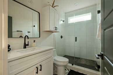 Bright modern bathroom with white vanity and sink, matte-black faucet and hardware, glass walk-in shower with narrow window, and patterned floor tiles.