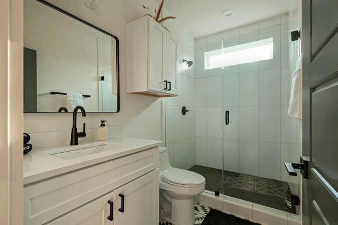 Bright modern bathroom with white vanity and sink, matte-black faucet and hardware, glass walk-in shower with narrow window, and patterned floor tiles.