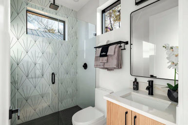 Modern sunlit bathroom with glass walk-in shower and geometric tile, black rain showerhead and fixtures, wood vanity with white countertop and mirror, towel rack with folded towels, and a white orchid by the window.