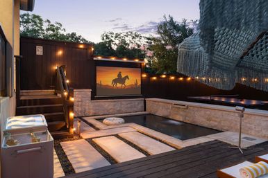 Cozy backyard patio at dusk with a small plunge pool, outdoor movie screen showing a cowboy silhouette, warm string lights, hammock fringe, stone stepping slabs and a cooler with drinks.