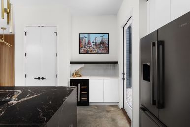 Contemporary home kitchen interior with a black marble island, white cabinetry, built-in wine fridge, black floating shelf and framed city artwork above, matte black French-door refrigerator beside a glass exterior door.
