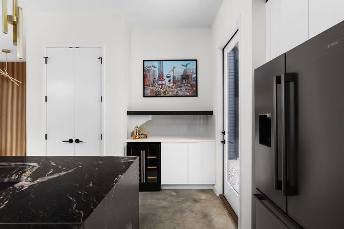 Contemporary home kitchen interior with a black marble island, white cabinetry, built-in wine fridge, black floating shelf and framed city artwork above, matte black French-door refrigerator beside a glass exterior door.