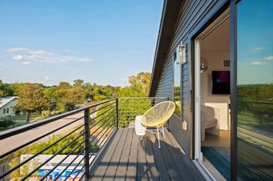 Sunny rooftop balcony with modern black metal railing and gray decking, yellow woven lounge chair and small white side table by a sliding glass door, overlooking a tree-lined suburban street and green treetops.