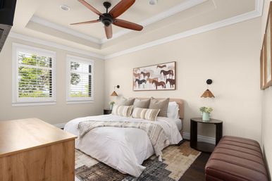 Sunlit neutral bedroom with tray ceiling and wooden ceiling fan, cozy bed dressed in layered beige linens and striped pillows, horse artwork above the headboard, plantation-shutter windows, pendant bedside lamps, round nightstands with potted flowers and a leather bench