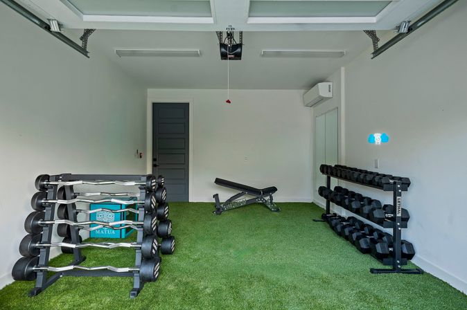 Home garage gym with green artificial turf under an open overhead door, racks of dumbbells and curl bars on either side, an adjustable weight bench centered, white walls and a wall-mounted air conditioner.