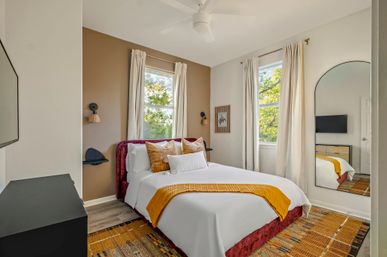 Cozy modern bedroom with burgundy velvet headboard, crisp white bedding and mustard yellow throw, two tall windows with cream curtains revealing a leafy outdoor view, arched wall mirror, ceiling fan and patterned rug.