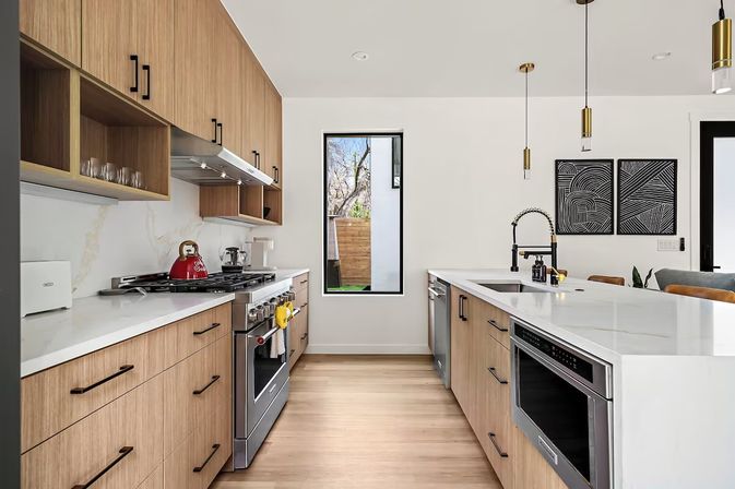 Bright modern open-concept kitchen with light wood cabinets, white quartz countertops, large island with black gooseneck faucet, stainless steel appliances, gas range with red kettle, brass pendant lights and narrow window.