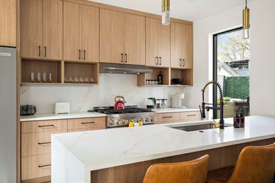 Bright modern kitchen with light wood cabinets, white marble-look quartz island and breakfast bar, stainless steel range and hood, black spring-coil pull-down faucet, gold pendant lights, brown leather stools, and a windowed view of a backyard.