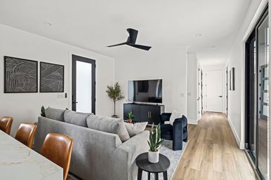 Bright modern open-plan living room with a gray sectional, navy armchair, black media console and TV, leather bar stools at a marble island, abstract black-and-white wall art, light hardwood floors, sliding glass door, potted plants and a black ceiling fan.