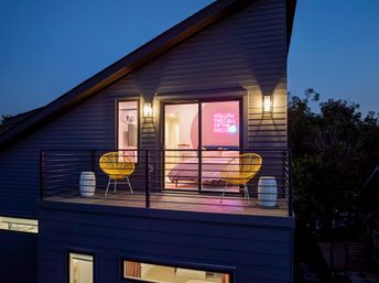 Contemporary two-story home at dusk with a second-story balcony, yellow woven chairs and ceramic side tables, sliding glass doors opening to a bedroom lit by a pink neon sign reading 'FOLLOW THE CALL OF THE DISCO' — playful, modern exterior scene.