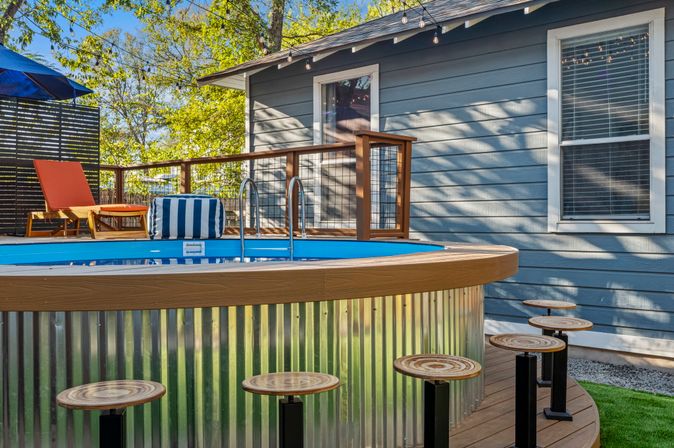Sunny backyard deck with round above-ground pool with corrugated metal exterior and wooden rim, ladder, striped cushion, orange lounge chairs, round wooden bar stools, and blue-gray house siding with string lights — inviting residential patio.