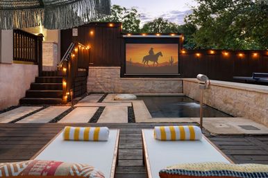 Cozy backyard patio at dusk with two striped-towel loungers facing a small pool and outdoor projector screen showing a cowboy silhouette, warm string lights and stone decking.