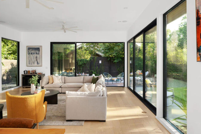 Bright, airy modern living room with a neutral sectional, mustard accent chair and coffee table, flooded with natural light from floor-to-ceiling sliding glass doors that open to a leafy backyard patio.