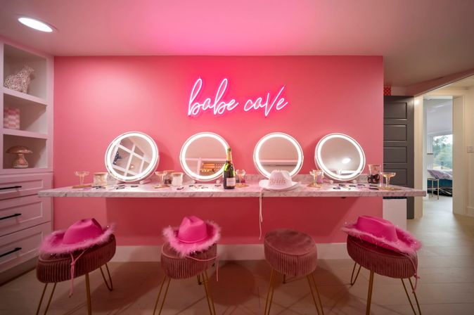 Bright pink bachelorette vanity with neon "babe cave" sign, four illuminated round mirrors above a marble counter holding champagne, a white "bride" hat and makeup, and four velvet stools with three pink feathered cowgirl hats.