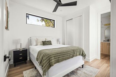 Bright modern minimalist bedroom with white bed, olive-green throw blanket, black nightstand and lamp, horizontal clerestory window, black ceiling fan, wood floors and an adjacent ensuite bathroom with wooden vanity.