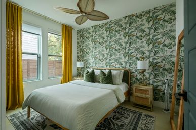 Bright tropical-style bedroom with botanical wallpaper, mustard-yellow curtains, twin windows, queen bed with green accent pillows, wooden ceiling fan and natural light.