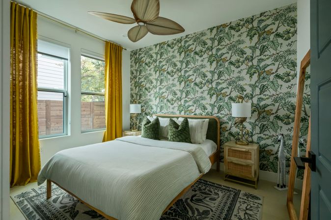 Bright tropical-style bedroom with botanical wallpaper, mustard-yellow curtains, twin windows, queen bed with green accent pillows, wooden ceiling fan and natural light.