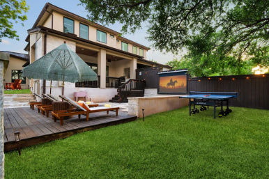 Spacious suburban two-story backyard patio at dusk with a wooden deck, fringed umbrella over chaise lounges, an outdoor movie screen showing a horseback silhouette, a ping-pong table on a green lawn, string lights and mature trees.