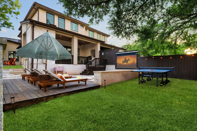 Spacious suburban two-story backyard patio at dusk with a wooden deck, fringed umbrella over chaise lounges, an outdoor movie screen showing a horseback silhouette, a ping-pong table on a green lawn, string lights and mature trees.