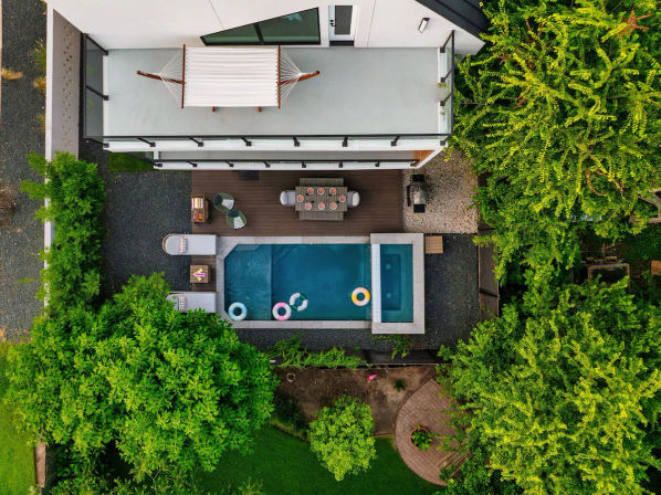 Aerial drone view of a contemporary backyard: rectangular pool with colorful float rings, wood deck with outdoor dining table and lounge seating, rooftop hammock, gravel paths and lush green trees surrounding the patio.
