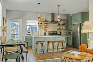 Bright open-concept kitchen and living area with mint-green cabinets, white subway tile backsplash, kitchen island with three wooden stools, stainless-steel appliances, rattan coffee table and cozy dining nook — modern coastal cottage vibe.