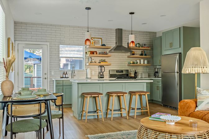 Bright open-concept kitchen and living area with mint-green cabinets, white subway tile backsplash, kitchen island with three wooden stools, stainless-steel appliances, rattan coffee table and cozy dining nook — modern coastal cottage vibe.
