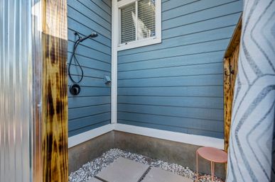 Cozy backyard outdoor shower in a blue-sided corner with a black hand shower, concrete stepping stones over white gravel, a small pink stool, and wood-and-metal privacy panels.