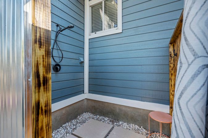 Cozy backyard outdoor shower in a blue-sided corner with a black hand shower, concrete stepping stones over white gravel, a small pink stool, and wood-and-metal privacy panels.