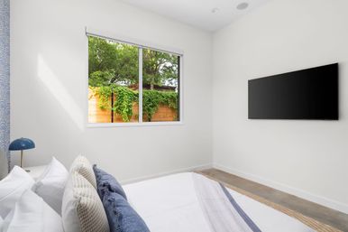 Bright modern minimalist bedroom with white walls, layered blue and neutral pillows on a neatly made bed, wall-mounted flat-screen TV, and a large window looking out to a wooden fence covered in green vines and trees.