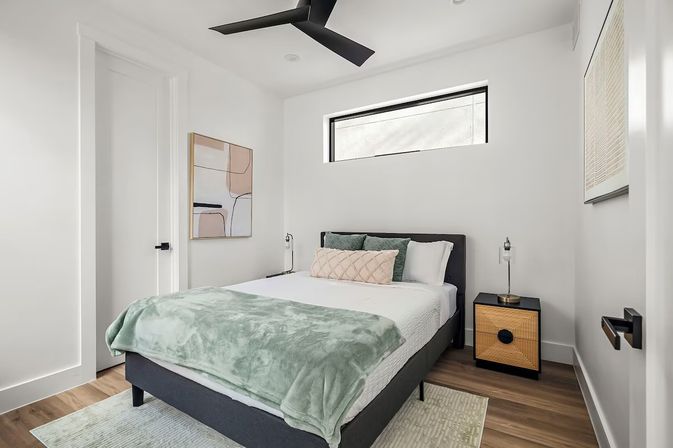 Sleek modern minimalist bedroom with dark gray upholstered bed, white linens and sage-green throw blanket, textured accent pillows, rattan-front nightstand, abstract wall art, narrow high window, black ceiling fan, and light wood floors.