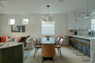 Modern open-plan dining and kitchen in a bright home — black dining table with tan leather chairs, sculptural white pendant, curved sofa with orange cushions, marble island and gold wire bar stools.