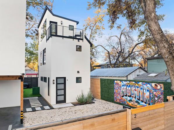 Contemporary three-story white micro‑home with rooftop deck, black trim and gravel courtyard, colorful 'Welcome to Austin, Texas' mural on the fence