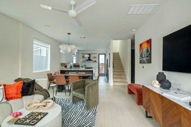 Light-filled modern open-concept living room and kitchen with pale wood floors, dining table and island, green velvet chair, zebra rug, staircase, and wall-mounted TV.