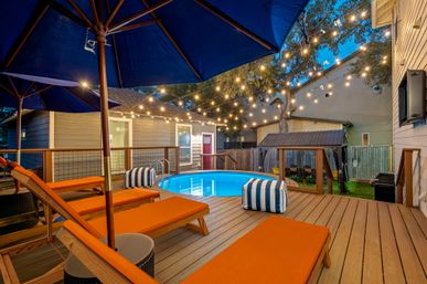 Cozy evening backyard deck with round above-ground pool, orange chaise lounges, blue umbrellas, striped poufs and string lights for relaxed outdoor entertaining.