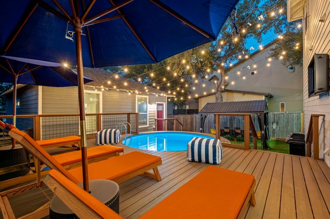 Cozy evening backyard deck with round above-ground pool, orange chaise lounges, blue umbrellas, striped poufs and string lights for relaxed outdoor entertaining.