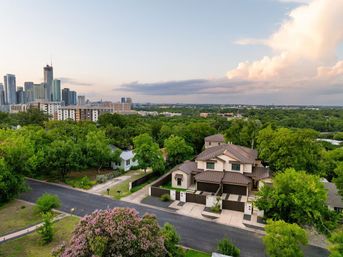 Aerial view of a residential neighborhood featuring a modern two-story townhouse, lush green trees and a distant city skyline beneath pastel sunset clouds.