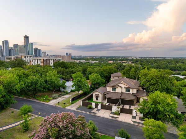 Aerial view of a residential neighborhood featuring a modern two-story townhouse, lush green trees and a distant city skyline beneath pastel sunset clouds.