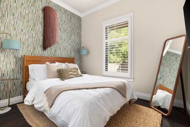 Cozy modern bedroom with mid-century wooden headboard, green patterned accent wall, white bedding, shuttered window, leaning full-length mirror, and woven jute rug.
