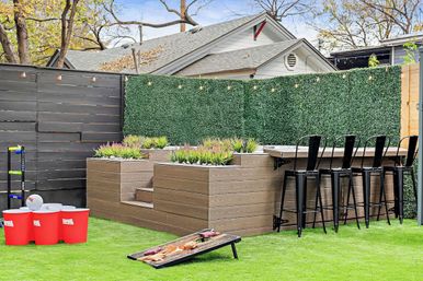 Residential backyard patio with artificial turf, tiered wood planters and green privacy hedge, string lights, raised outdoor bar with black metal stools, and lawn games including cornhole and oversized red cup toss