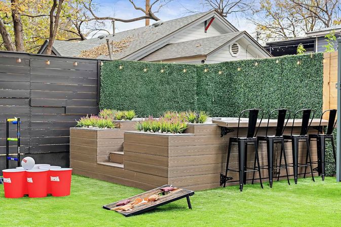 Residential backyard patio with artificial turf, tiered wood planters and green privacy hedge, string lights, raised outdoor bar with black metal stools, and lawn games including cornhole and oversized red cup toss