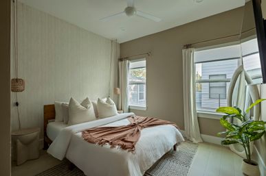 Cozy sunlit modern bedroom with white linens, plush cream pillows, rust-colored fringed throw, wooden headboard, hanging woven pendant lights, ceiling fan, large mirror, potted plant, neutral curtains and textured rug
